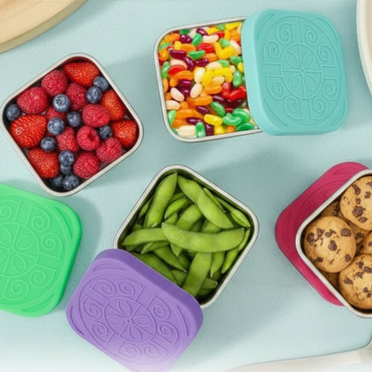 Colorful snack containers with berries, jelly beans, snap peas, and cookies on a light blue surface.